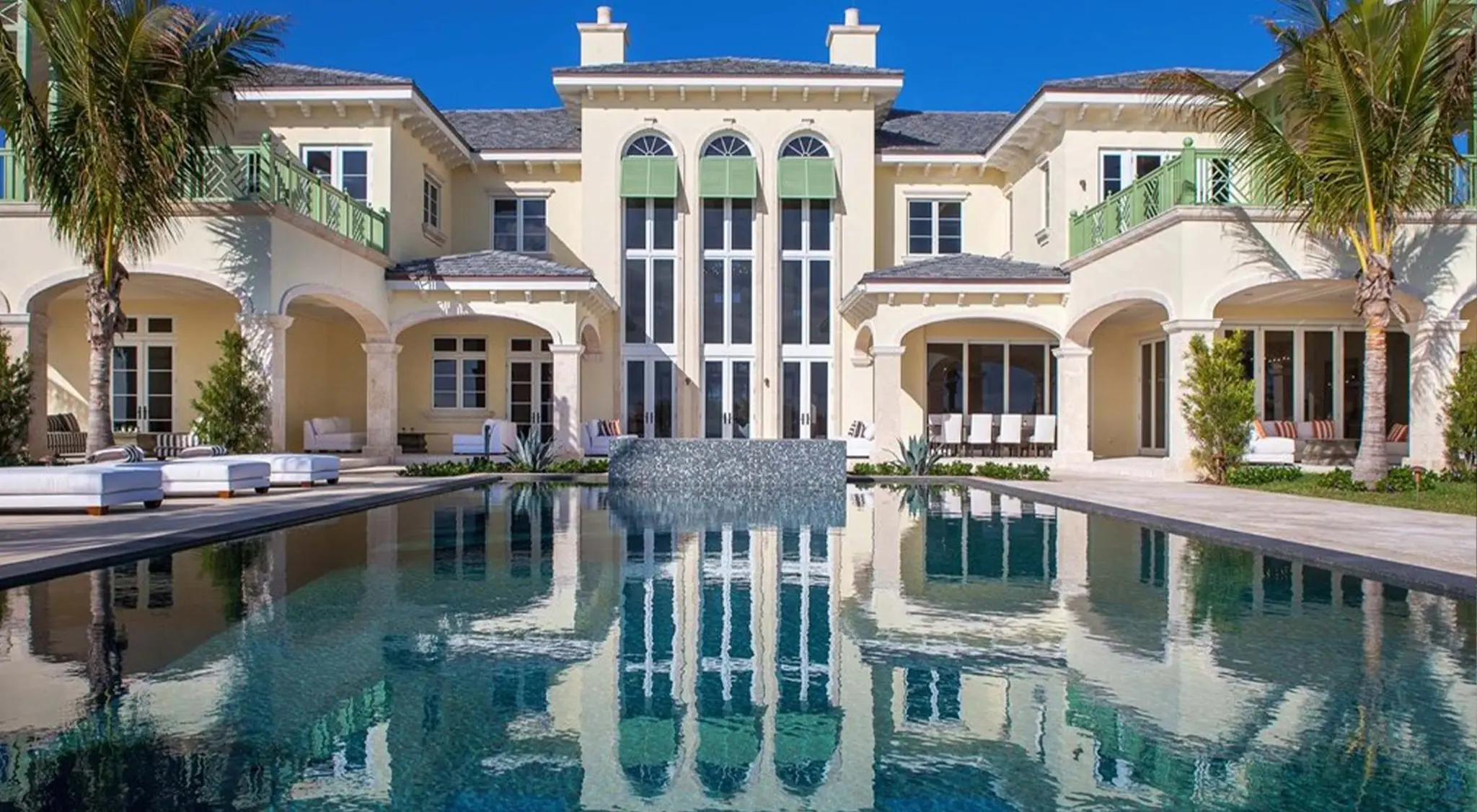 A finished cream stucco estate with green balcony railings behind a long reflecting pool and palms, the last frame of the build film (illustrative, generated).
